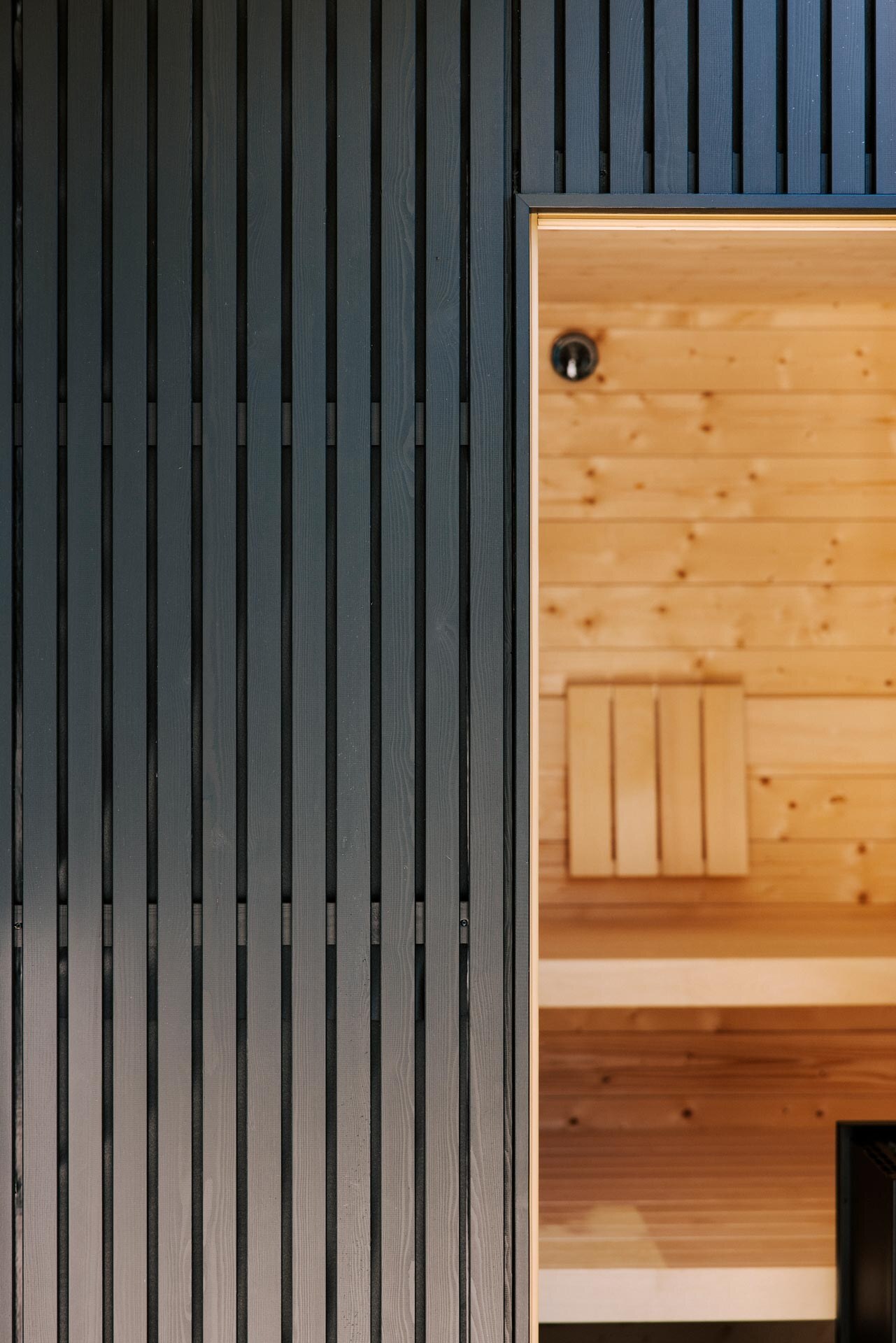 Outdoorsauna mit minimalistischer, schwarzer Außenverkleidung aus Holzlatten und hellem Fichtenholz im Inneren.
