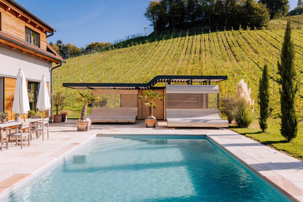 Individuelle Outdoor-Sauna mit wellenförmigem Sonnenliegen, integriertem Sichtschutz und großem Pool in einem Garten am Weinberg.