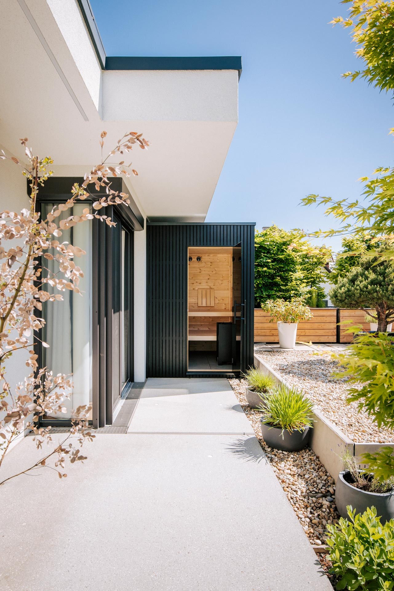 Schwarz verkleidete Outdoorsauna auf einer Terrasse in einem begrünten Steingarten.