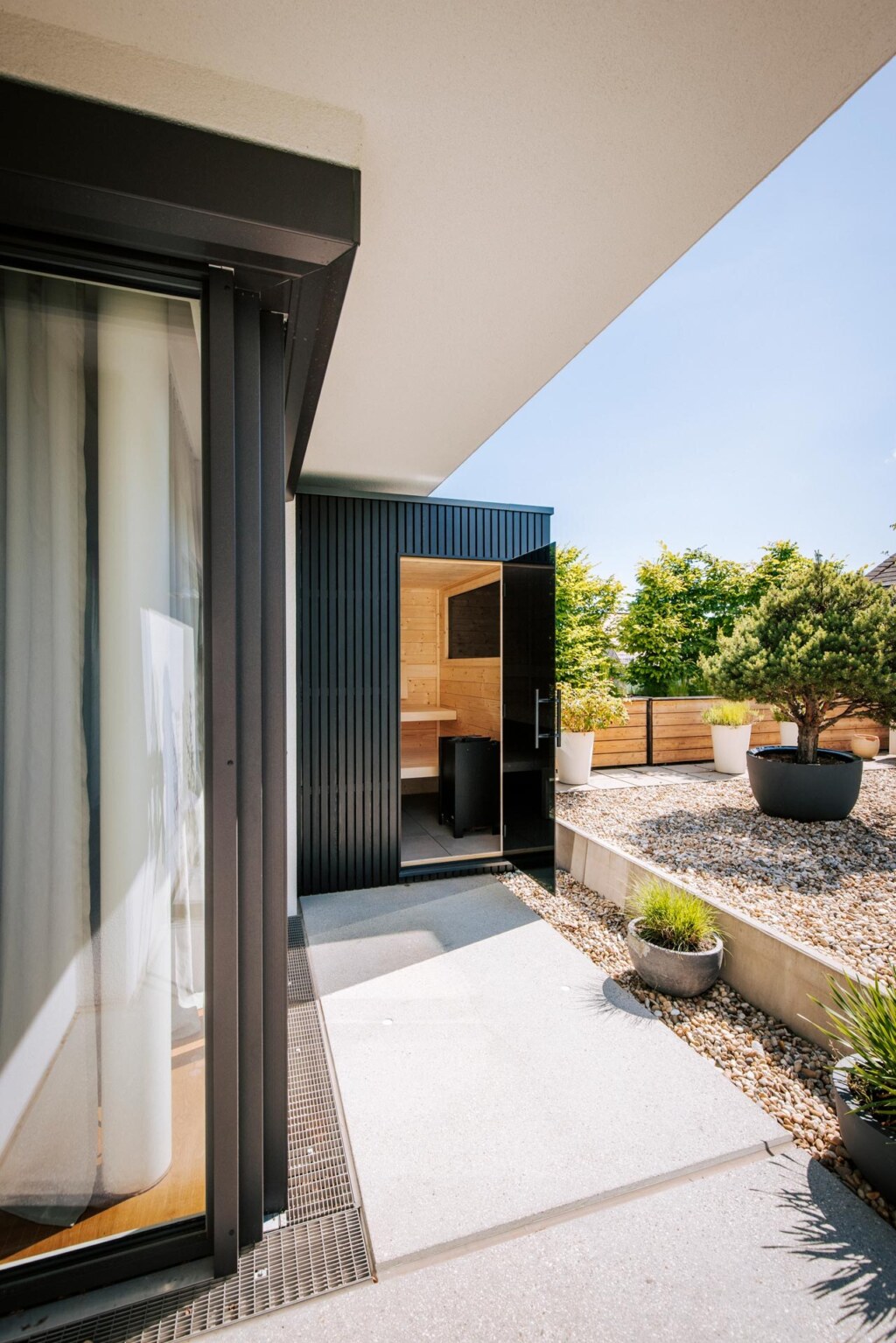 Kleine moderne Designsauna mit schwarzer Holzverkleidung unter einem Balkon im Steingarten.