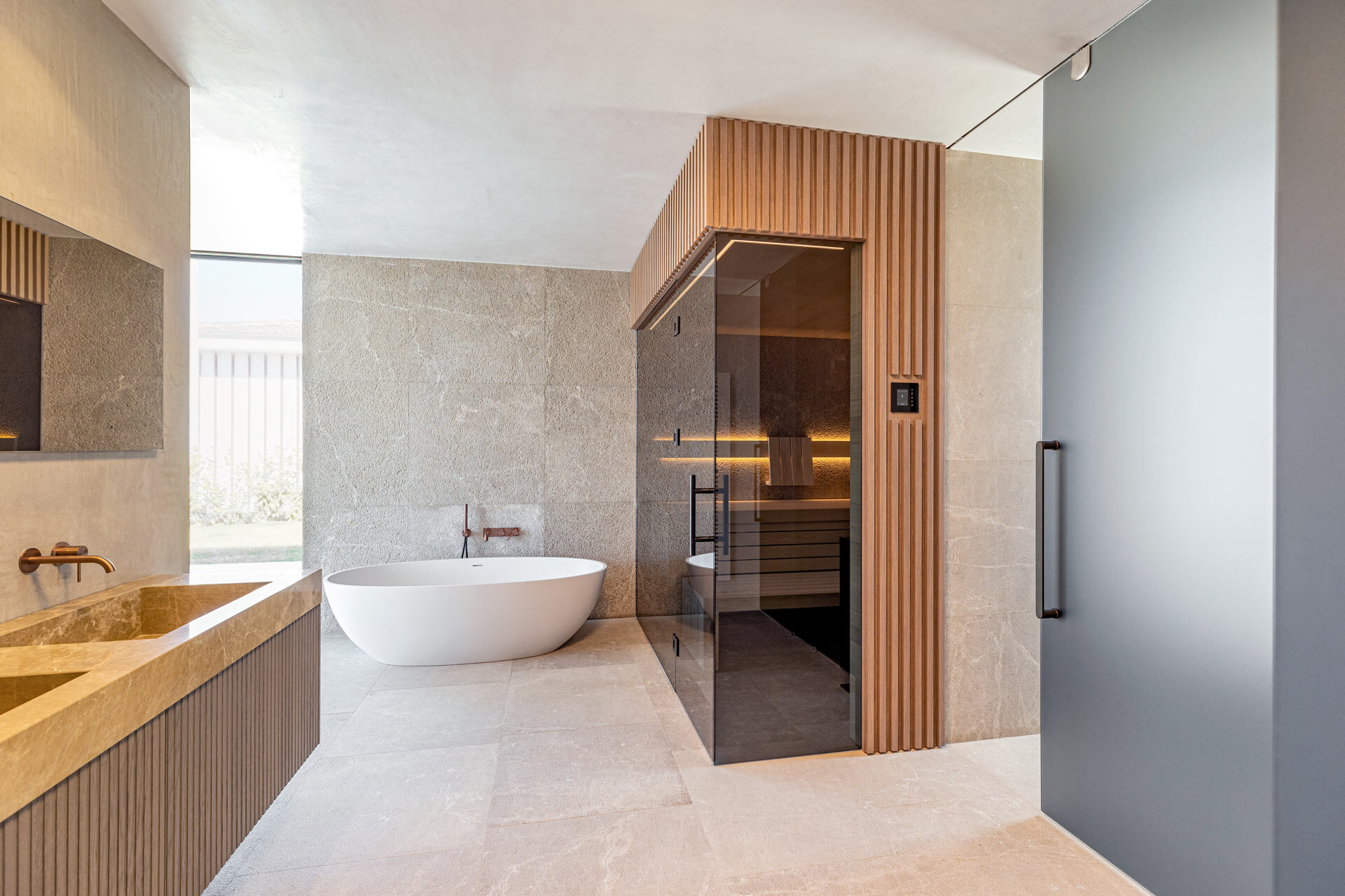 Individuell gestaltete Designsauna mit getönter Frontverglasung, einer Außenverlleidung aus getönten Holzleisten, in einem luxuriösen Badezimmer aus Marmor und Naturstein mit freistehender Badewanne und Waschtisch.