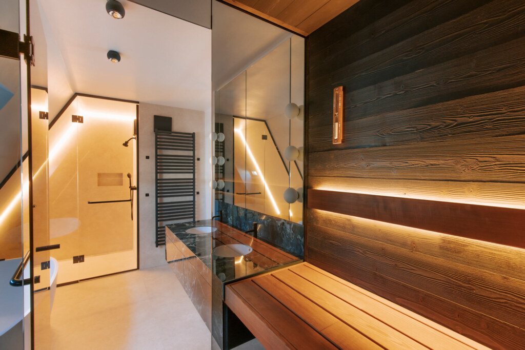 Ausblick aus einer Sauna aus dunklem Thermofichtenholz auf eine beleuchtete Dusche in einem modernen Badezimmer im Dachgeschoss.