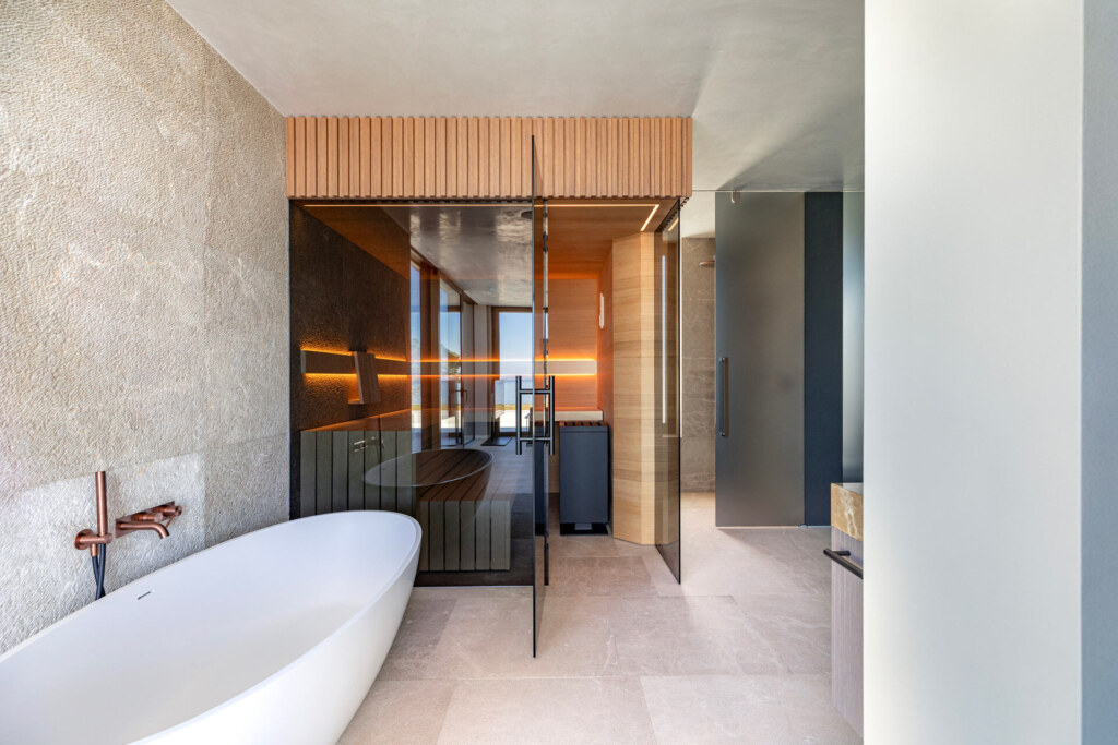 Moderne Sauna mit getönter Frontverglasung, außen verkleidet mit Holzleisten, innen ausgeführt in gemaserten, astfreien Eichenholzpaneelen in einem Badezimmer aus Marmor und Naturstein mit freistehender Badewanne.