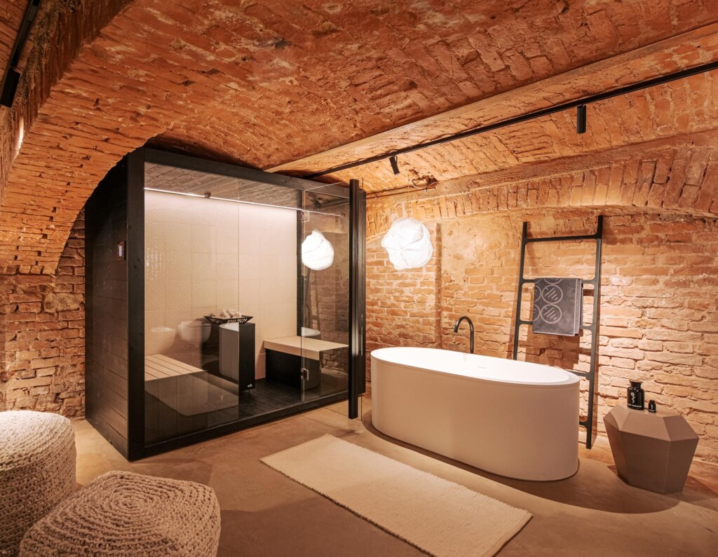 Kompakte Sauna und eine freistehende Badewanne im Gewölbekeller des Deisl Showroom im Formdepot Wien. Die Sauna ist in schwarzem Lärchenholz ausgeführt, mit einer großen Glasfront ausgestattet und innen an der Rückwand in weißen Keramikfliesen verkleidet.