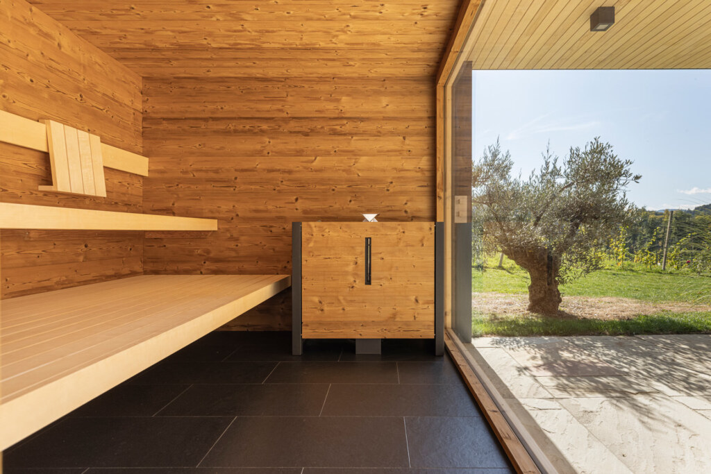 Sauna von Deisl aus Thermo-Fichte mit einem großem Panoramafenster und Blick auf einen mediterranen Garten mit Olivenbaum.