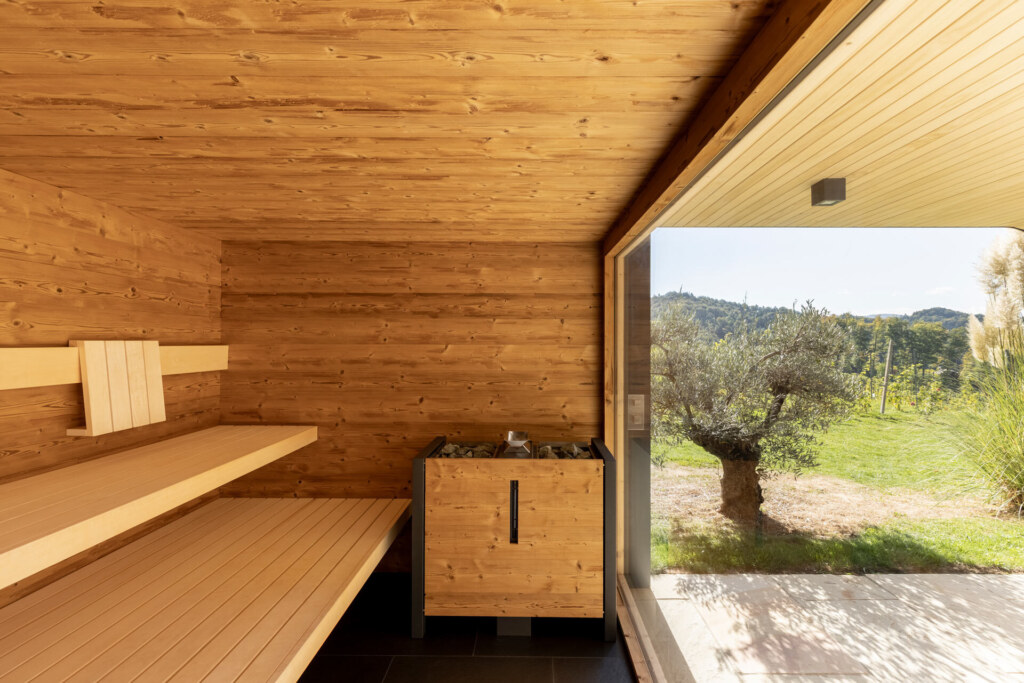 Sauna von Deisl aus Thermo-Fichte mit einem großem Panoramafenster und Blick auf die Terrasse und einen mediterranen Garten mit Olivenbaum.