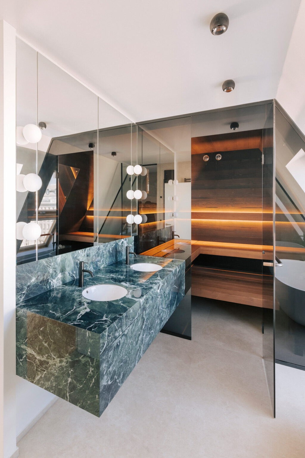 Maßgeschneiderte Sauna in individuellem Design aus dunklen Hölzern, schwarzer Glasfront und mit direkt anschließendem Waschtisch aus grünem Marmor, harmonisch unter einer Dachschräge eines modernen Badezimmers eingebaut.