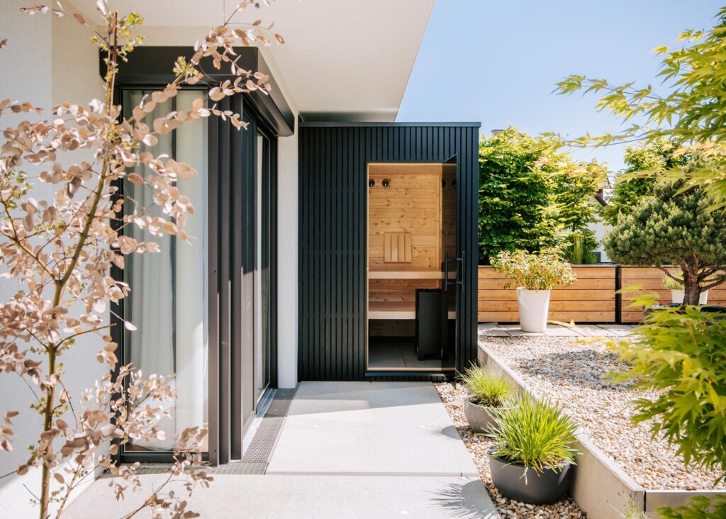 Schwarz verkleidete Outdoorsauna auf einer Terrasse in einem begrünten Steingarten.