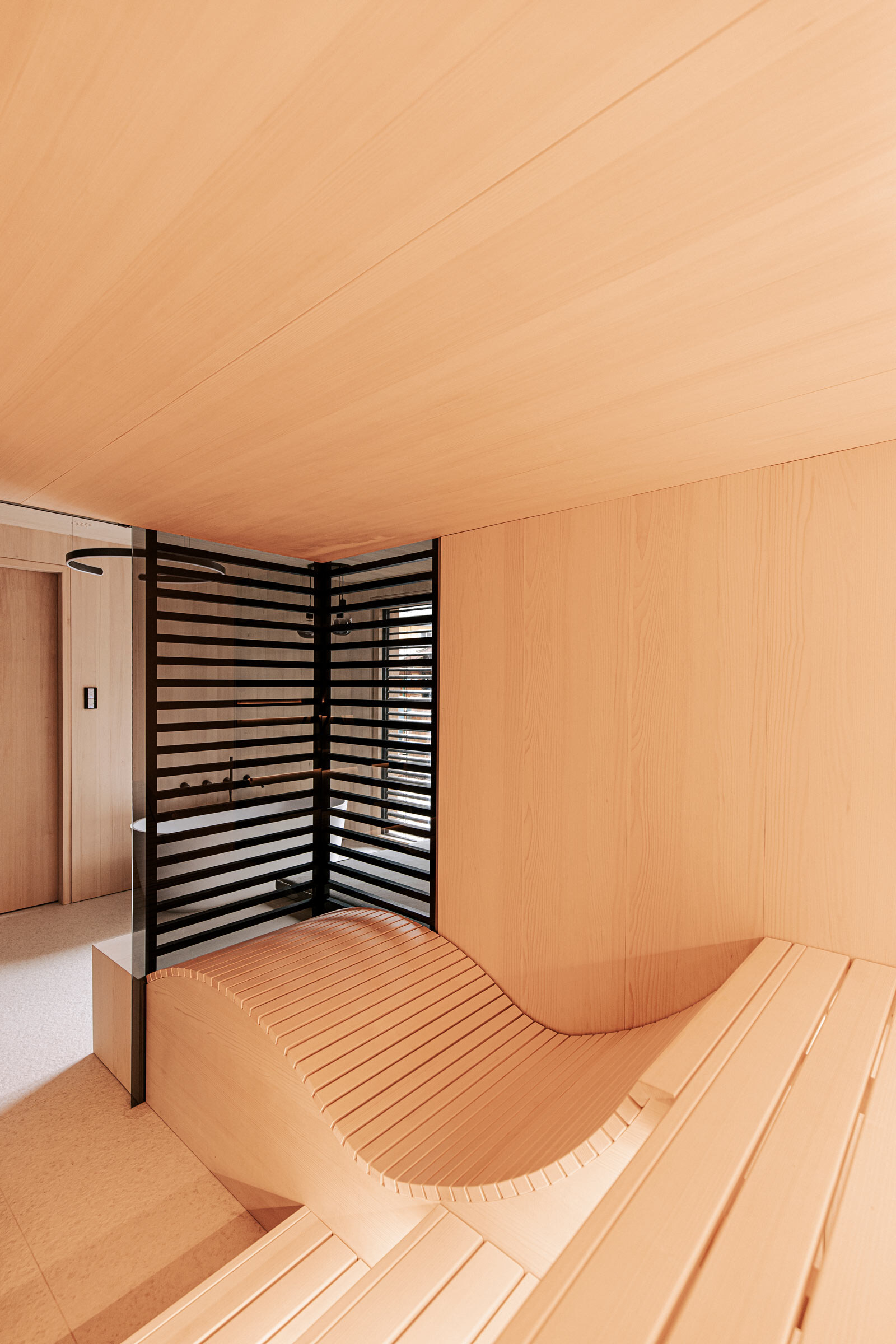 Ausblick aus einer minimalisitschen Sauna in eine modernes Badezimmer, ausgeführt in hellem gesaegtem Tannenholz, mit ergonomischer Saunaliege und raumhoher Verkleidung aus schwarz getoenten Holzstreben als Sichtschutz.