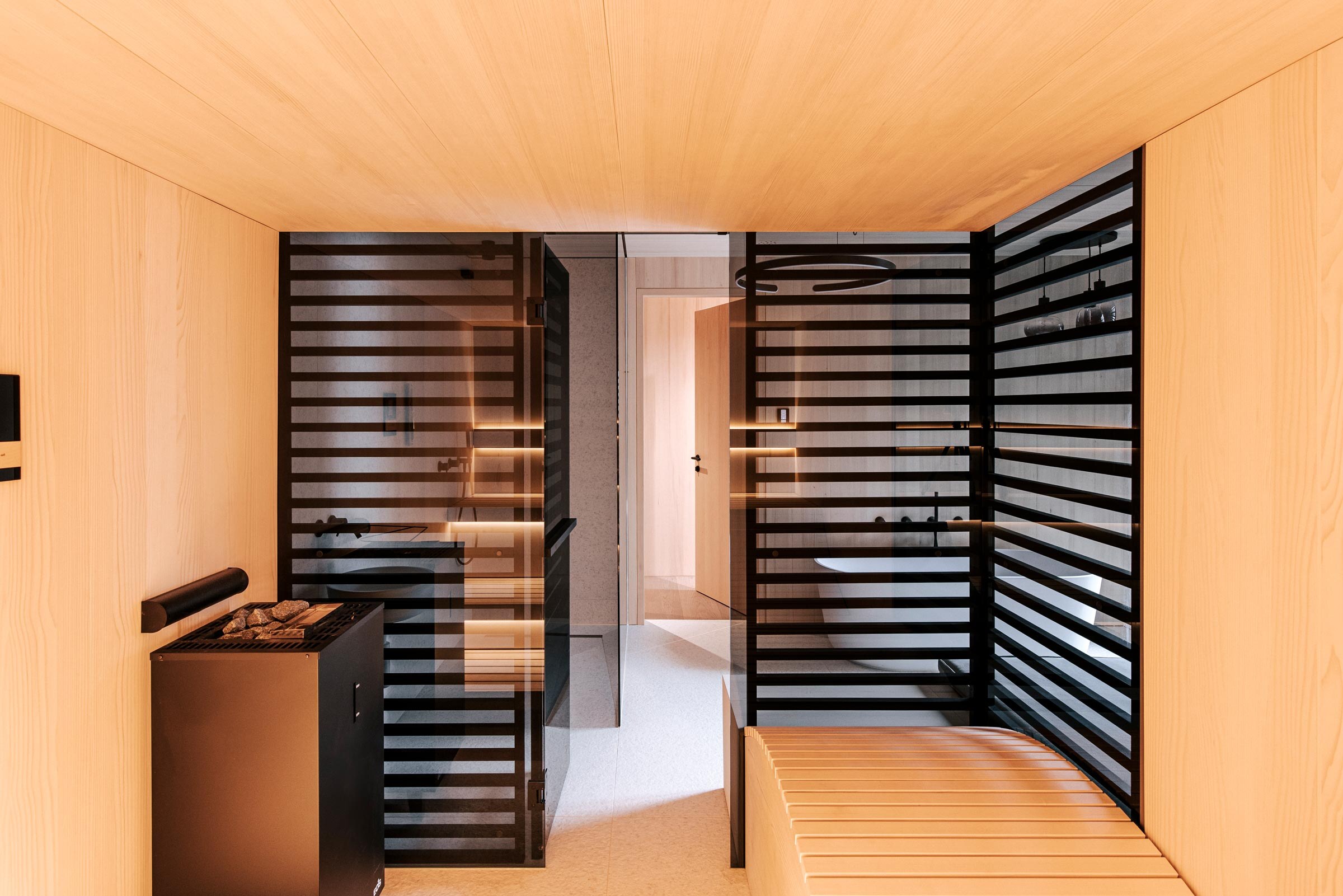 Ausblick aus der Sauna in das Badezimmer im Scandi Stil, ausgefuehrt in hellen gesaegtem Tannenholz, mit ergonomischer Saunaliege und raumhoher Verkleidung aus schwarz getoenten Holzstreben als Sichtschutz.