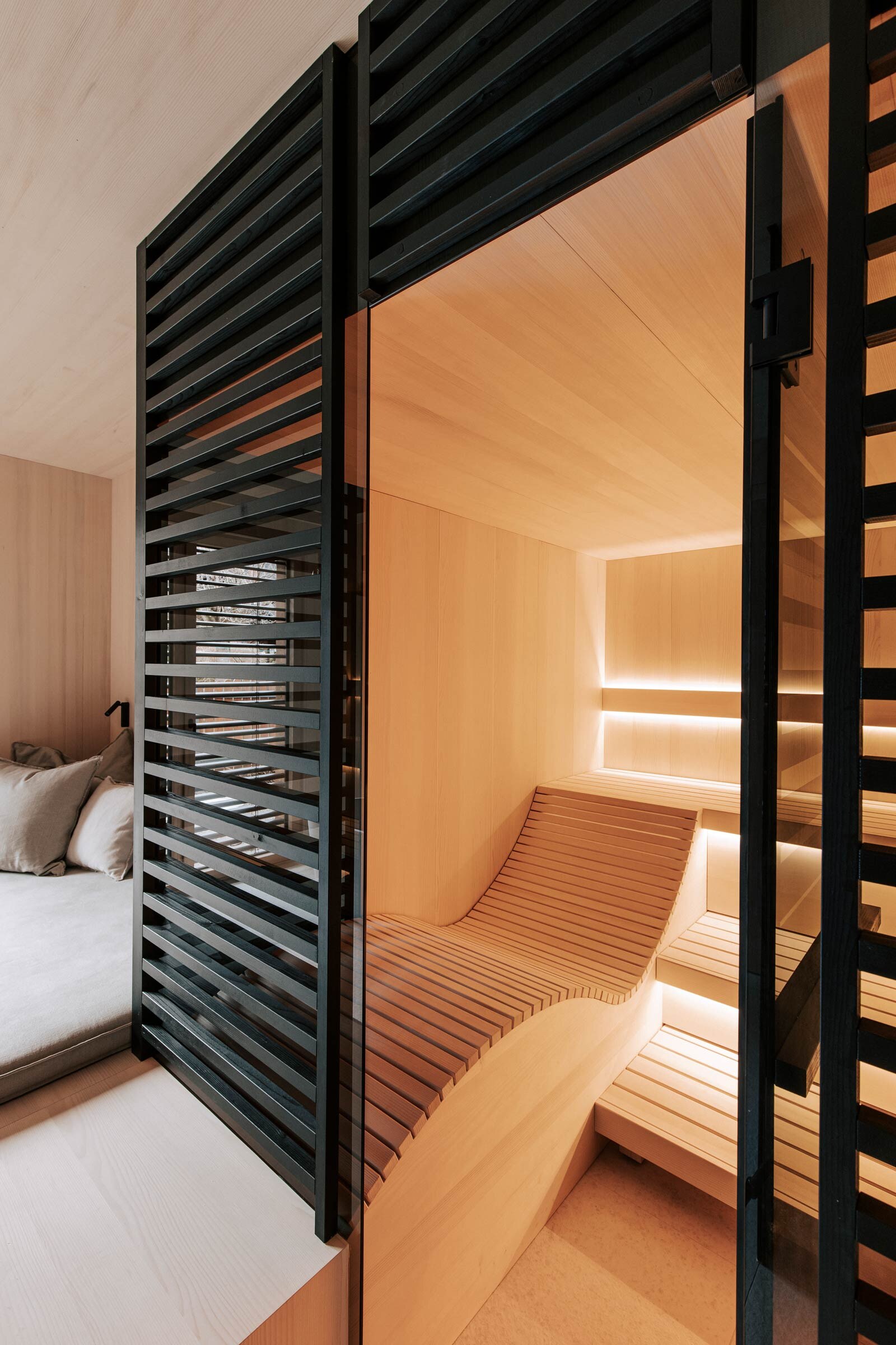 Massgeschneiderte Sauna mit ergonomischer Liege aus Espenholz, getoenter Panorama-Glasfront, sowie raumhoher Verkleidung aus schwarz getoenten Holzstreben als Sichtschutz.