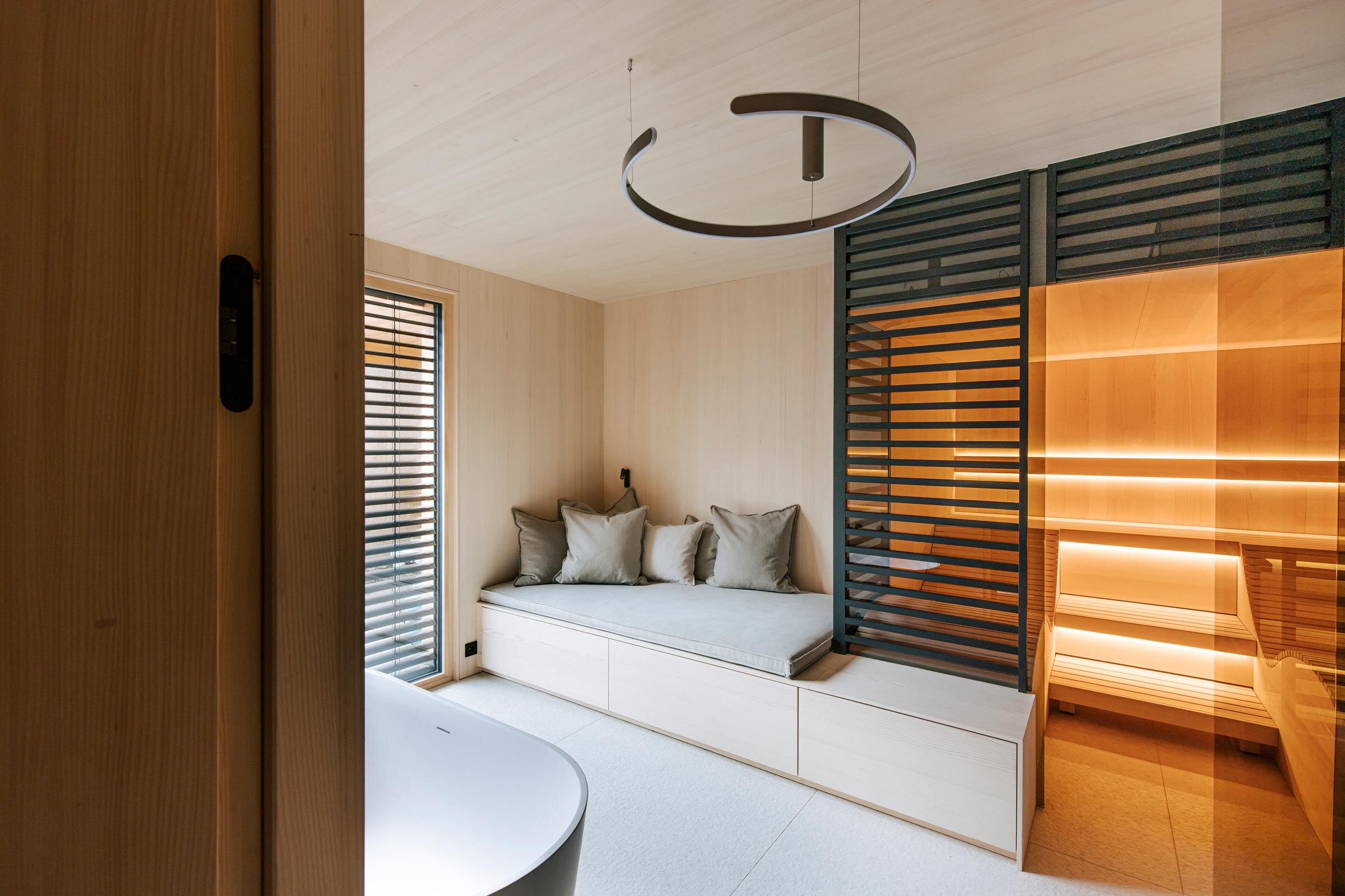 Beleuchtete Design-Sauna von Deisl, ausgeführt in Tannenholz mit Sichtschutz aus schwarz getoenten Holzstreben, anschliessendem Daybed mit Verstaumoeglichkeit, sowie freistehende Badewanne im Badezimmer mit heller Holzverkleidung.