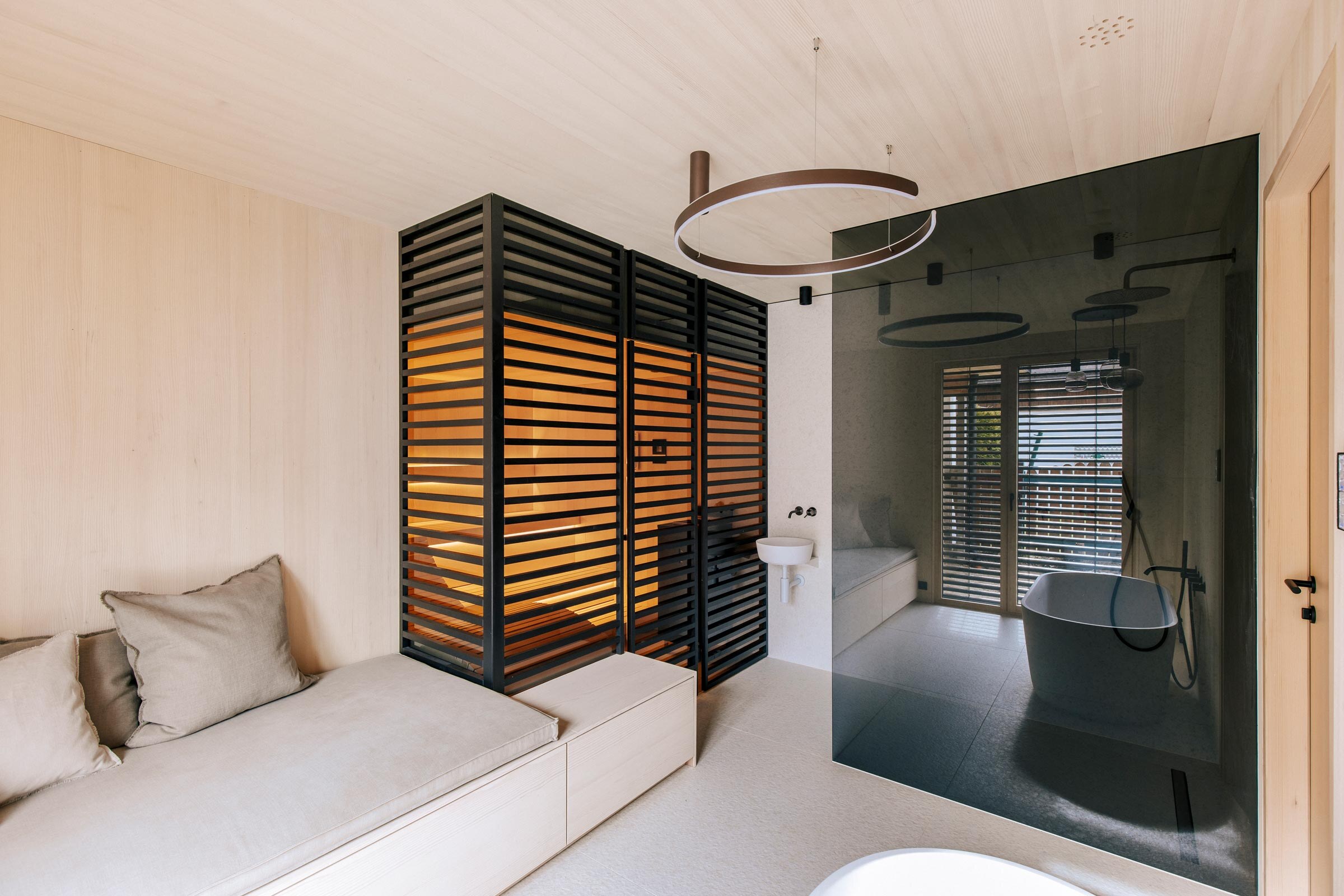 Designsauna der Marke Deisl, im Badezimmer aus Tannenholz, mit getoenter Panorama-Glasfront, sowie raumhoher Verkleidung aus schwarz getoenten Holzstreben als Sichtschutz, anschliessendem Daybed, freistehender Badewanne und bodengleicher Dusche.