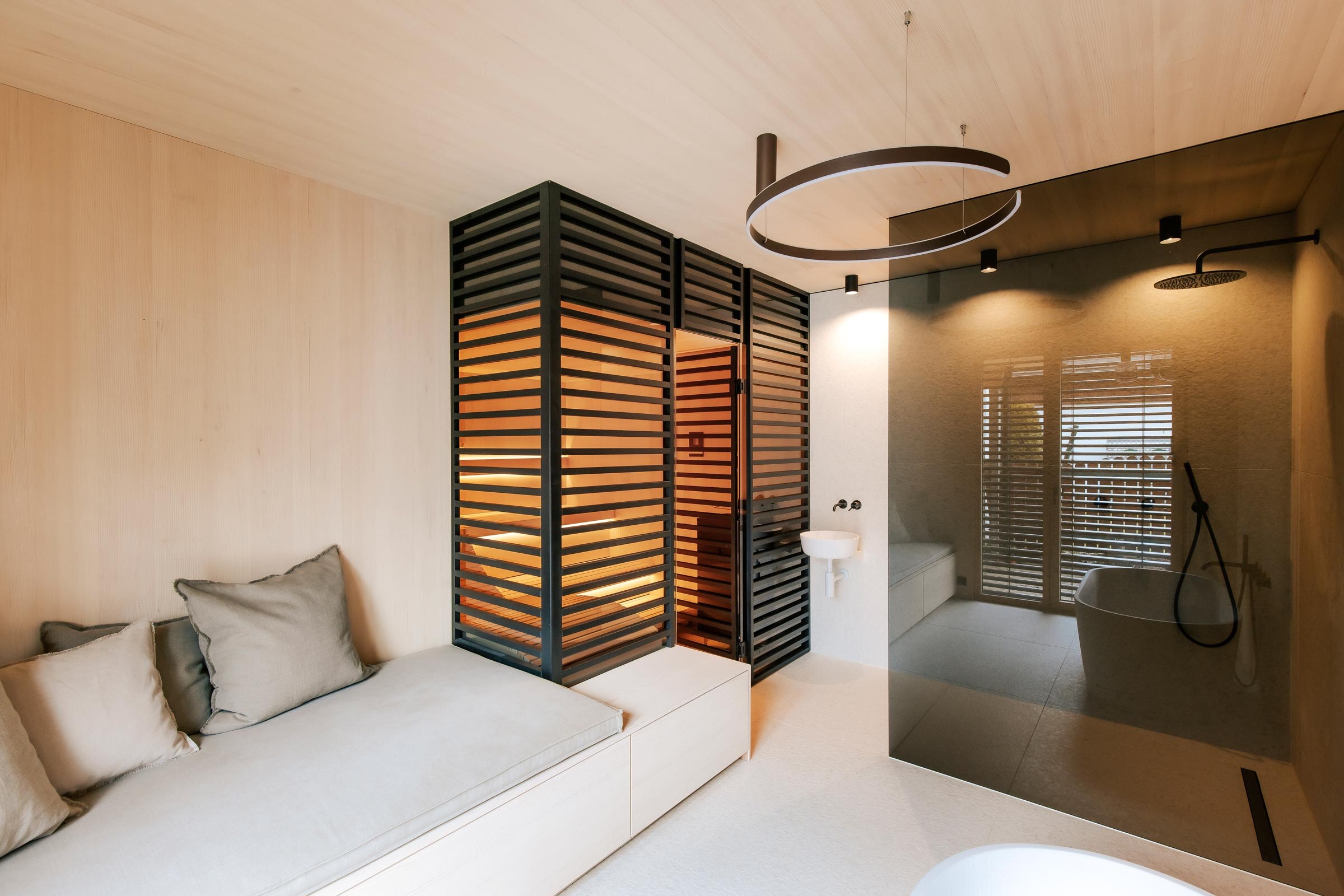 Massgeschneiderte Deisl Sauna im Badezimmer mit getoenter Panorama-Glasfront, sowie raumhoher Verkleidung aus schwarz getoenten Holzstreben als Sichtschutz, anschliessendem Daybed und bodengleicher Dusche.