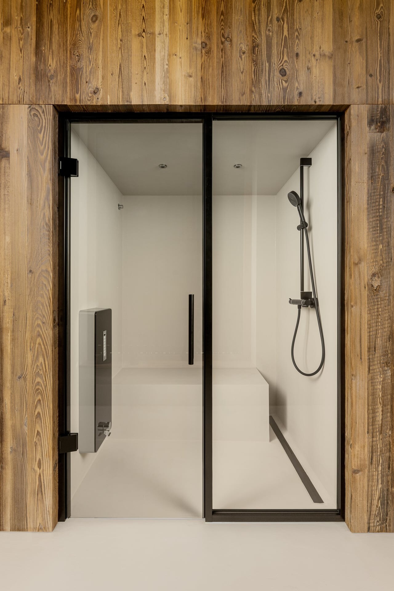 Elegante Dampfdusche in mininmalistisch-alpinem Design aus Mikrozement mit Glasfront, schwarzen Armaturen und Holzverkleidung im Chalet-Badezimmer.