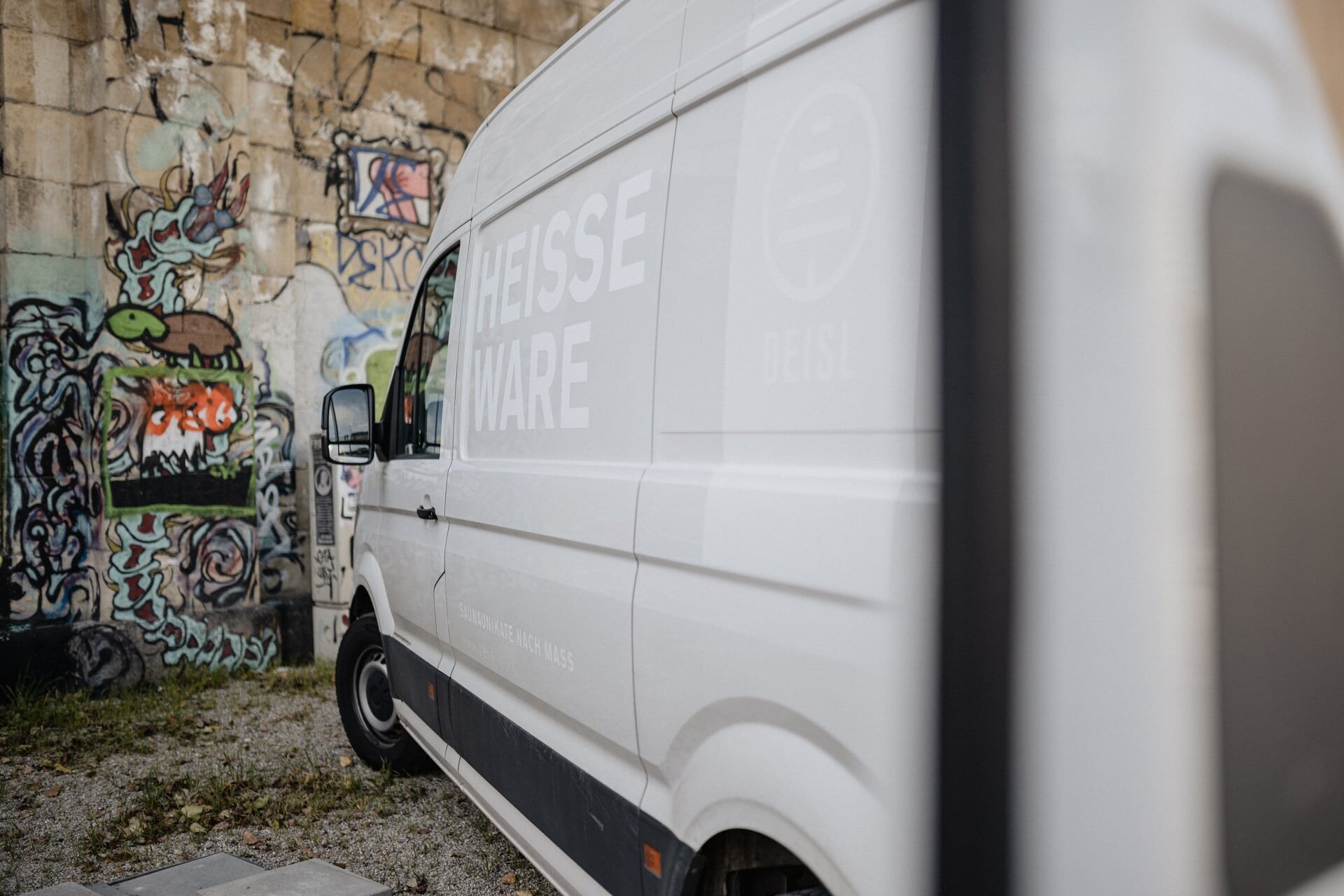 Ein weißer Deisl Sauna-Montagebus mit der Aufschrift Heisse Ware parkend vor einer urbanen Graffiti-Wand.