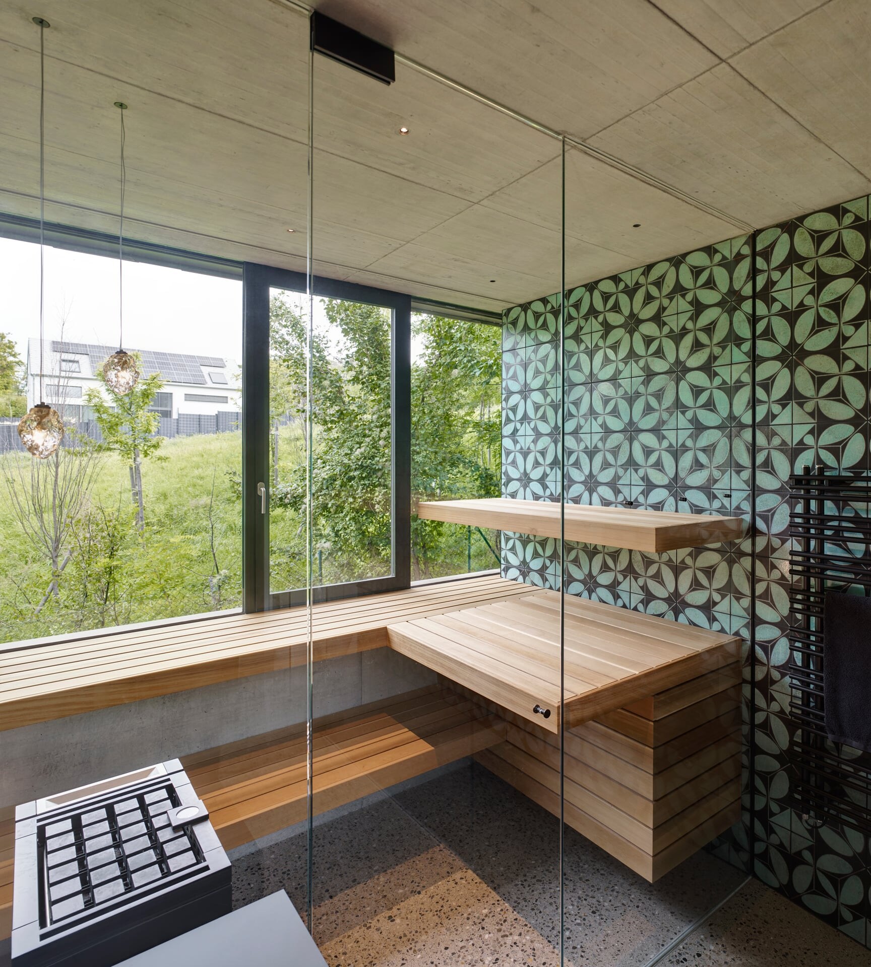 Design-Sauna aus Glas mit ornamentaler Fliesenwand, Bänken aus Zedernholz und Fenster mit Blick ins Grüne.
