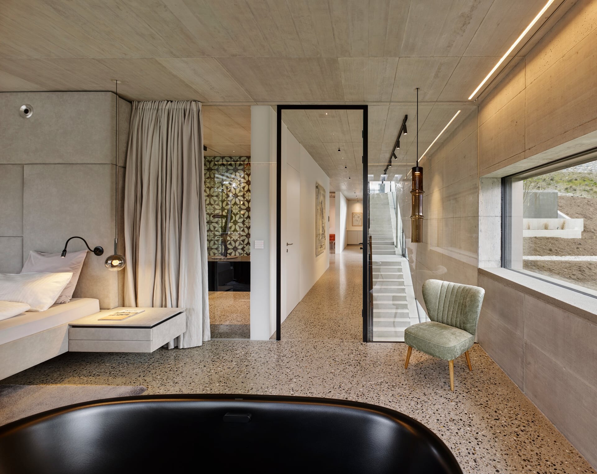 Modernes Schalfzimmer aus Sichtbeton mit Terrazzoboden und freistehender, schwarzer Badewanne.